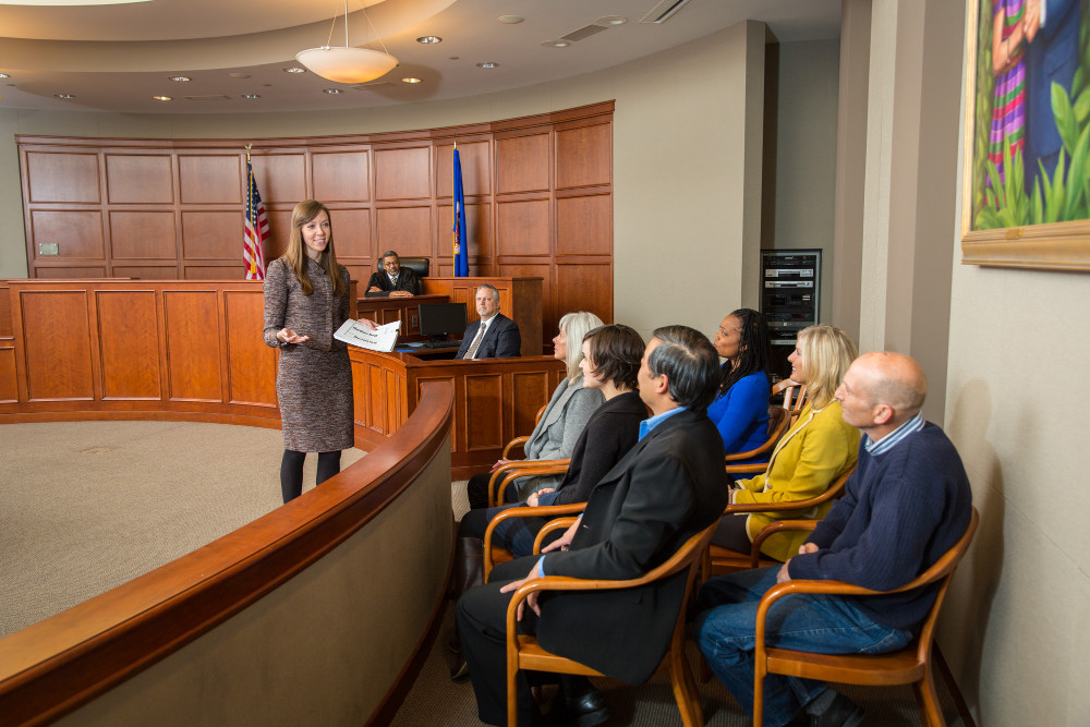 The image shows a courtroom scene with a Erin Conway Johnsen standing and addressing a jury. The jury is seated to her right, and there is a judge sitting behind a desk.