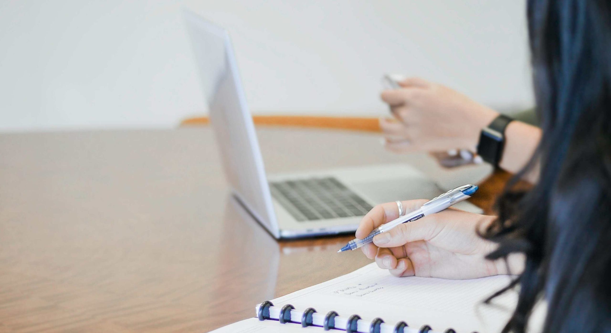 In the image, two people are sitting at a table. One is writing in a notebook, and the other is using a laptop and a phone