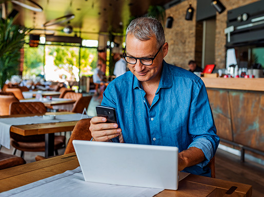 A man sitting at a table in a restaurant with a laptop and cell phone in hand.