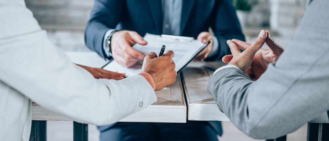 Three men in suits sitting at a table signing paperwork